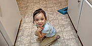 Melody joined the competition — help win amazing prizes! child, smiling, kitchen, floor, tile_floor, crouching, furniture, table, chair, cabinet, counter, cup, person, indoor, young_child, casual_clothing, hair, expression, domestic, playful