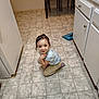 child, smiling, kitchen, floor, tile_floor, crouching, furniture, table, chair, cabinet, counter, cup, person, indoor, young_child, casual_clothing, hair, expression, domestic, playful