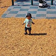 Miles is registered to the contest to win money with this photo: toddler, child, playground, wood_chips, toy_car, bucket_hat, mickey_mouse, shorts, shoes, sunlight, shadow, outdoor, play_area, person, smiling, daytime, recreation, fun, park, casual_clothing