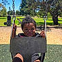 Miles is registered to the contest to win money with this photo: toddler, child, swing, playground, smiling, happy, outdoor, park, grass, trees, blue_sky, sunny, play, fun, black_swing, chains, curly_hair, daytime, nature, person