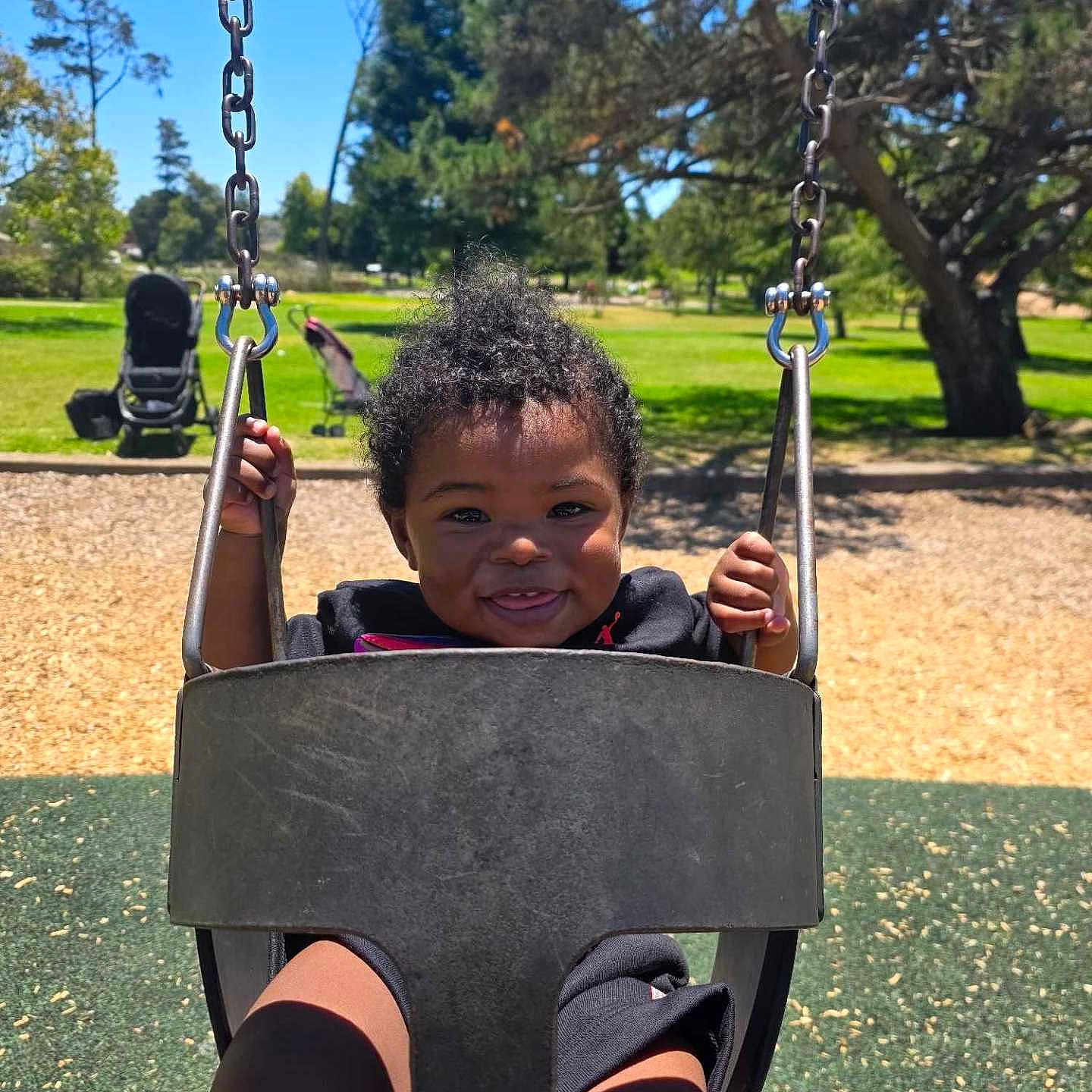 Miles is registered to the contest to win money with this photo: black_swing, blue_sky, chains, child, curly_hair, daytime, fun, grass, happy, nature, outdoor, park, person, play, playground, smiling, sunny, swing, toddler, trees