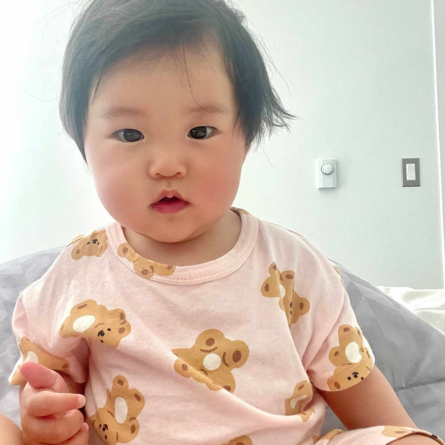 Iris joined the competition — help win amazing prizes! baby, bed, black_hair, blanket, child, curly_hair, cute, face, hand, indoor, innocent_expression, light_switch, pink_clothing, portrait, short_sleeves, sitting, teddy_bear_pattern, thermostat, toddler, white_wall