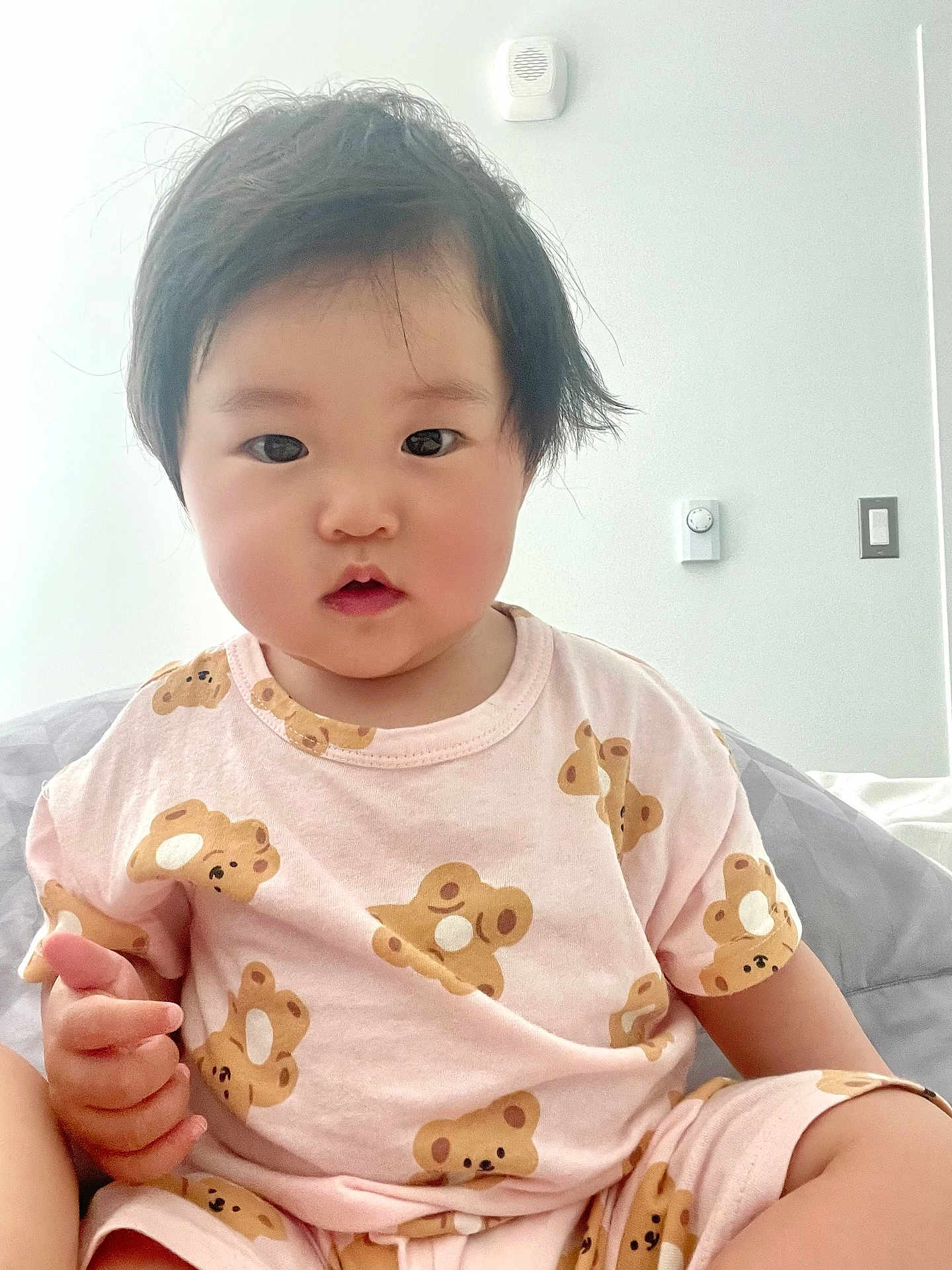 Iris joined the competition — help win amazing prizes! toddler, child, baby, pink_clothing, teddy_bear_pattern, short_sleeves, indoor, white_wall, light_switch, thermostat, bed, blanket, black_hair, curly_hair, cute, portrait, sitting, hand, face, innocent_expression
