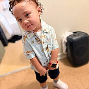 Jesiah is registered to the contest to win money with this photo: child, toddler, portrait, curly_hair, smiling, indoor, standing, fashion, patterned_shirt, shorts, sneakers, smartwatch, suitcase, bedroom, tile_floor, full_body, close_up, casual_clothing, playful, home