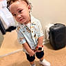 child, toddler, portrait, curly_hair, smiling, indoor, standing, fashion, patterned_shirt, shorts, sneakers, smartwatch, suitcase, bedroom, tile_floor, full_body, close_up, casual_clothing, playful, home