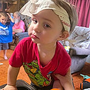 Aiden Gehringer is registered to the contest to win money with this photo: toddler, child, headband, red_shirt, teenage_mutant_ninja_turtles, living_room, furniture, couch, curtains, carpet, toy, person, hand, orange_toy, background, indoor, casual_clothing, boy, adult, family