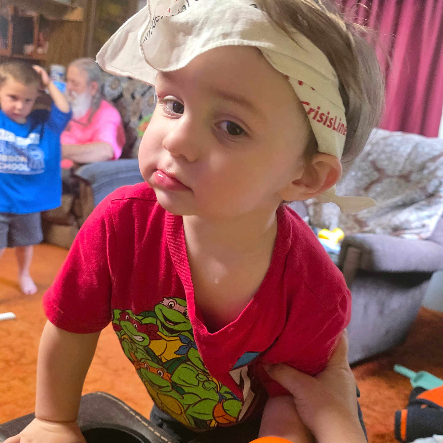 Aiden Gehringer is registered to the contest to win money with this photo: toddler, child, headband, red_shirt, teenage_mutant_ninja_turtles, living_room, furniture, couch, curtains, carpet, toy, person, hand, orange_toy, background, indoor, casual_clothing, boy, adult, family