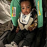 baby, child, car_seat, sneakers, bib, plaid_shirt, clothing, person, indoor, seat_belt, infant, footwear, curly_hair, looking, portrait, cute, expression, carrying_device, fabric, small_child