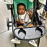 Prince is registered to the contest to win money with this photo: baby, stroller, child, sneakers, bib, green_seat, black_stroller, person, indoor, curly_hair, face, hands, footwear, casual_clothing, baby_boy, seatbelt, wheel, floor, toy_accessory, toddler
