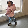 toddler, child, indoor, window, pink_vest, jeans, black_sneakers, curly_hair, pigtails, floor, wall, casual_clothing, looking_back, young_child, portrait, standing, natural_light, curious, small_child, cute