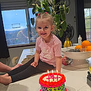 Lillian joined the competition — help win amazing prizes! banana, birthday_cake, blonde_hair, cake, candles, celebration, child, countertop, curtains, fruit_bowl, happy, indoor, kitchen, orange, person, pink_shirt, plant, smiling, table, window