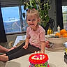 banana, birthday_cake, blonde_hair, cake, candles, celebration, child, countertop, curtains, fruit_bowl, happy, indoor, kitchen, orange, person, pink_shirt, plant, smiling, table, window