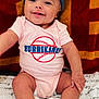 baby, child, smiling, headband, bow, pink_onesie, baseball_logo, blanket, script_text, sitting, happy, infant, cute, skin, feet, hands, face, portrait, indoor, colorful_background