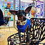 animal, bandana, blue_jacket, blurred_background, cafe, chair, colorful, dog, ears, floor, glass_door, indoor, light, modern_chair, neon_sign, pet, shop, small_dog, standing, window