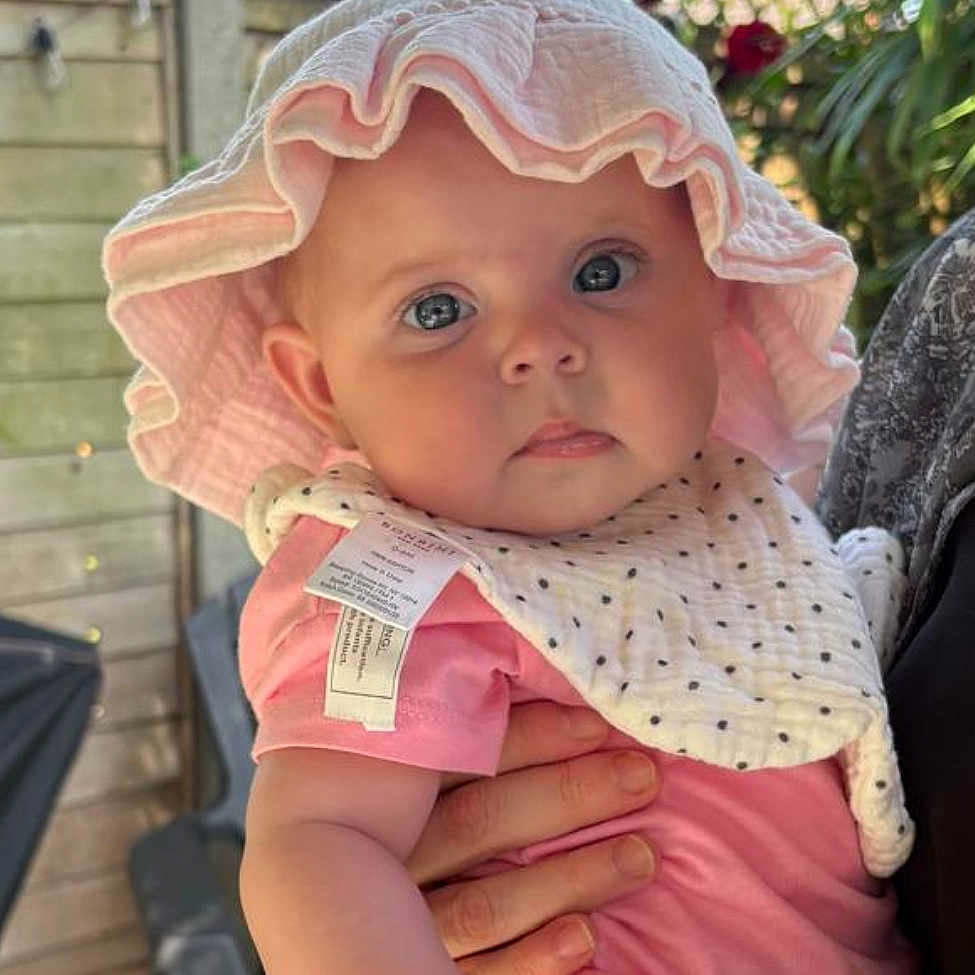 Madison Iris joined the competition — help win amazing prizes! baby, bib, child, cute, daylight, eyes, face, fence, flowers, hand, holding, infant, nature, outdoor, person, pink_clothing, portrait, skin, summer, sunhat