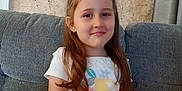 Prayla is registered to the contest to win money with this photo: child, girl, smiling, sitting, couch, white_tshirt, yellow_shorts, hair_accessory, indoor, portrait, happy, wall_art, painting, home_interior, curtain, furniture, casual_clothing, young, person, relaxed