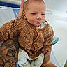 baby, infant, knitted_sweater, tattoo, hand, person, clothing, blue_towel, white_background, cute, newborn, portrait, holding, expression, sitting, cozy, cute_face, small_hands, patterned_pants, indoor