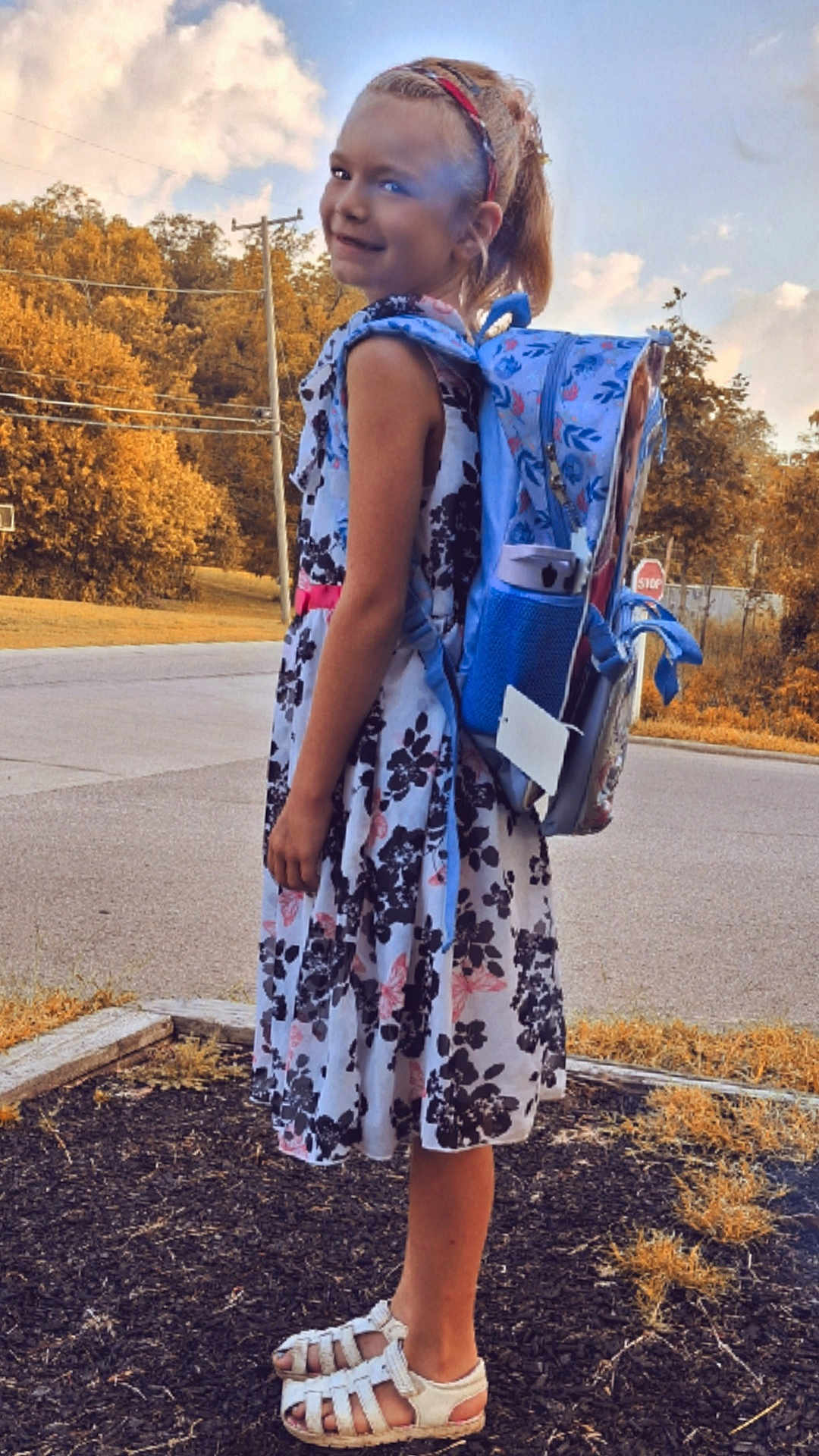 Kensley is registered to the contest to win money with this photo: child, girl, backpack, dress, sandals, outdoor, road, trees, stop_sign, sidewalk, nature, smiling, standing, headband, summer, happy, person, footwear, sky, daytime