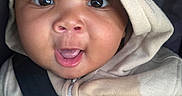 Prince is registered to the contest to win money with this photo: baby, hoodie, seatbelt, closeup, child, infant, smiling, happy, face, clothing, hand, blanket, portrait, cute, expression, indoors, person, young_child, black_hair, warm_clothing