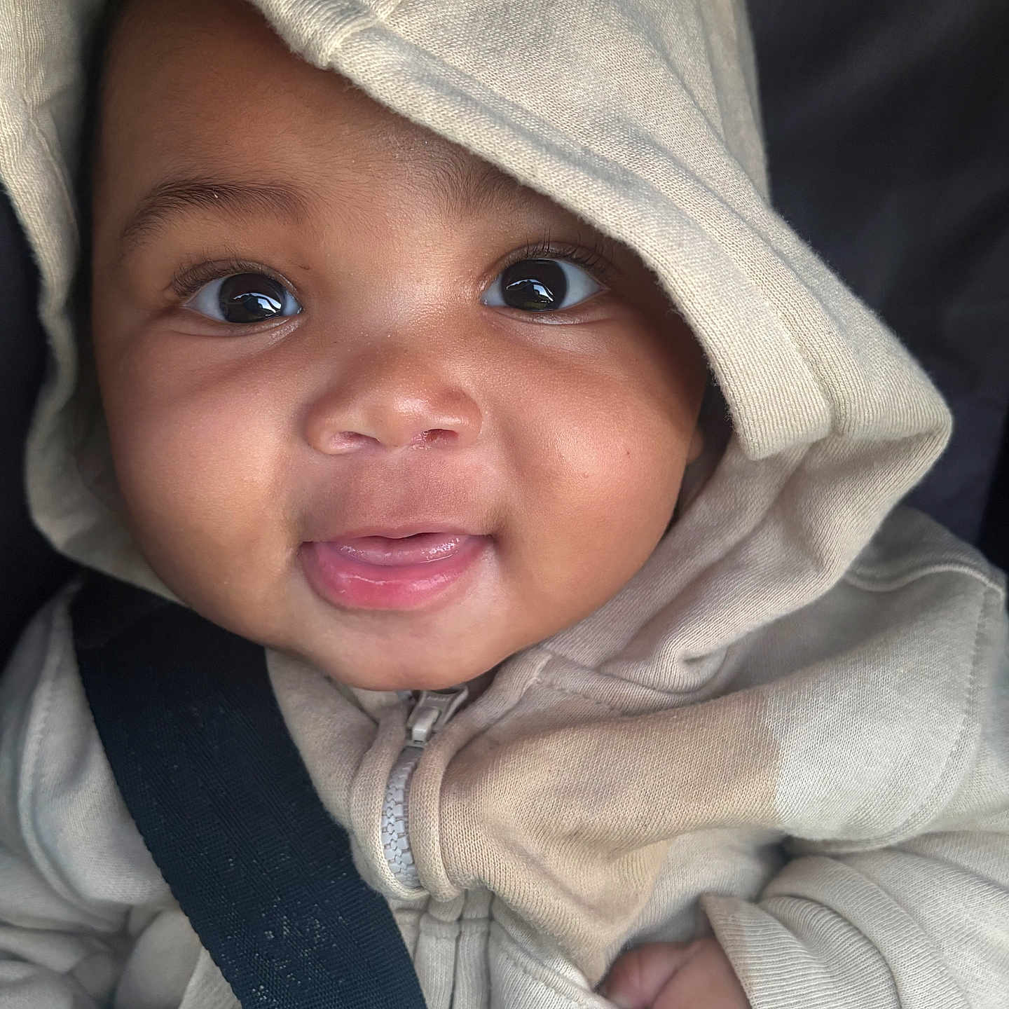 Prince is registered to the contest to win money with this photo: baby, child, hoodie, seatbelt, car_seat, smile, face, cute, infant, clothing, closeup, portrait, dark_skin, happy, young_child, person, casual_wear, warm, indoors, expression
