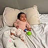 Melody is registered to the contest to win money with this photo: baby, infant, child, portrait, bed, bedding, pillow, blanket, sleeping_area, indoors, bedroom, pink_clothing, patterned_pants, socks, toy, rattle, serious_expression, sitting, cozy, home