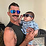 adult, baby, baby_playpen, blue_shirt, casual_wear, child, family, furniture, hand, happy, holding, indoor, man, mustache, patterned_clothing, portrait, short_hair, smile, sunglasses, tattoo