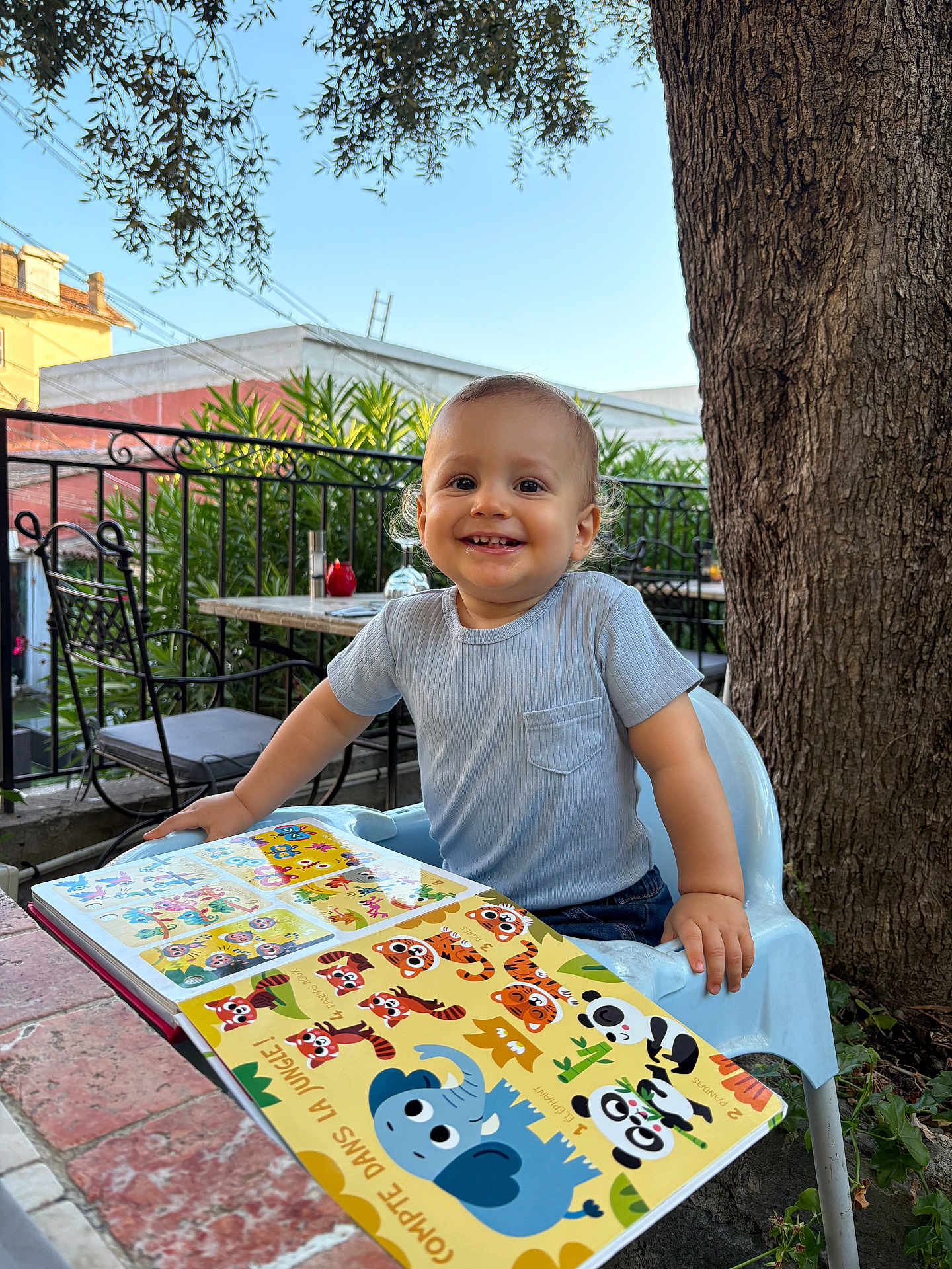 Gabriel participe au concours pour gagner de l'argent avec cette photo : book, child, clothing, curly_hair, daylight, face, furniture, garden, greenery, happy, outdoor, person, plastic_chair, playful, reading, short_sleeve, smiling, table, toddler, tree