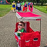 Brantley is registered to the contest to win money with this photo: child, boy, wagon, red, sunglasses, smiling, park, path, people, group, cape, superhero, outdoor, trees, grass, playground, canopy, walking, daytime, cloudy