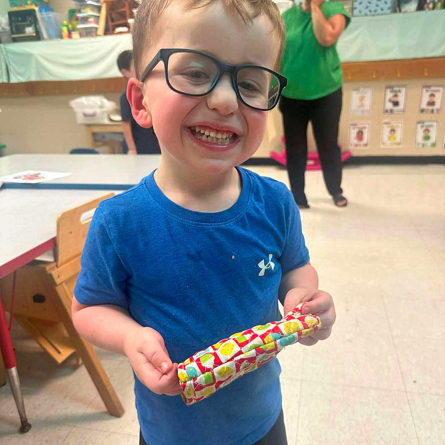 Brantley is registered to the contest to win money with this photo: child, boy, glasses, smiling, blue_shirt, shorts, classroom, adult, educational_materials, table, chair, floor, toy, person, indoor, happy, people, background, furniture, colorful