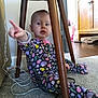 baby, child, pajamas, colorful, pointing, wooden_table, carpet, floor, indoor, person, furniture, curious, expression, sitting, cable, wall, cabinet, footwear, home, playful