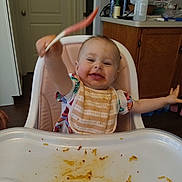 Layla joined the competition — help win amazing prizes! baby, high_chair, spoon, bib, messy, food, kitchen, cabinet, happy, smiling, child, person, indoor, tray, tableware, plastic, clothing, face, arm, wood_floor