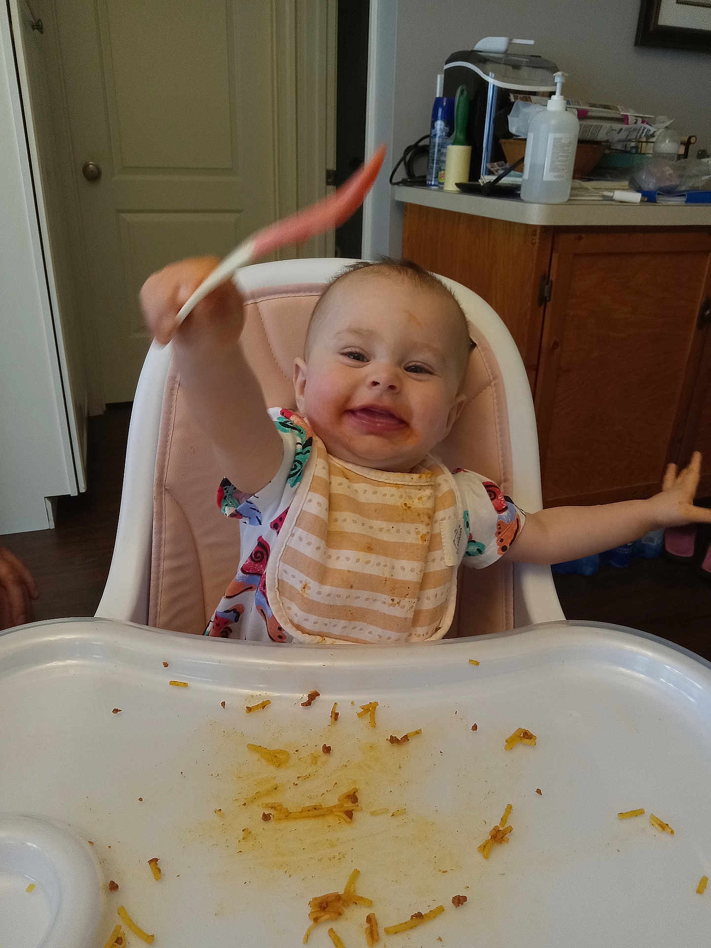 Layla joined the competition — help win amazing prizes! baby, high_chair, spoon, bib, messy, food, kitchen, cabinet, happy, smiling, child, person, indoor, tray, tableware, plastic, clothing, face, arm, wood_floor