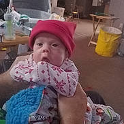 Daniel is registered to the contest to win money with this photo: baby, infant, red_hat, pajamas, blanket, hand, indoor, person, face, carseat, table, bottle, living_room, yellow_basket, furniture, cozy, holding, home, cute, child