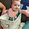 baby, infant, bib, fabric_basket, couch, quilt, arm, hand, smiling, indoor, cozy, blanket, patterned, textile, person, child, cute, happy, seated, closeup