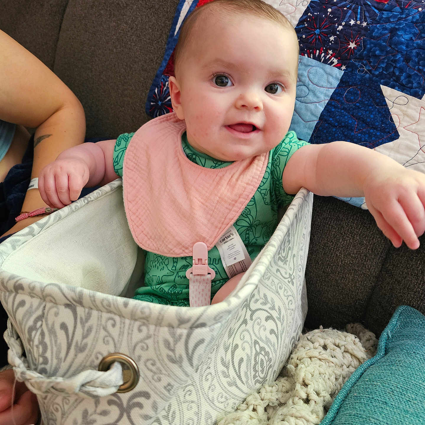 Everly is registered to the contest to win money with this photo: baby, infant, bib, fabric_basket, couch, quilt, arm, hand, smiling, indoor, cozy, blanket, patterned, textile, person, child, cute, happy, seated, closeup