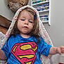 toddler, child, superman_shirt, blanket, indoor, bookshelf, books, knitted_blanket, blue_shirt, face, hair_clip, furniture, person, young_child, casual_clothing, seated, brown_hair, expression, home, curious