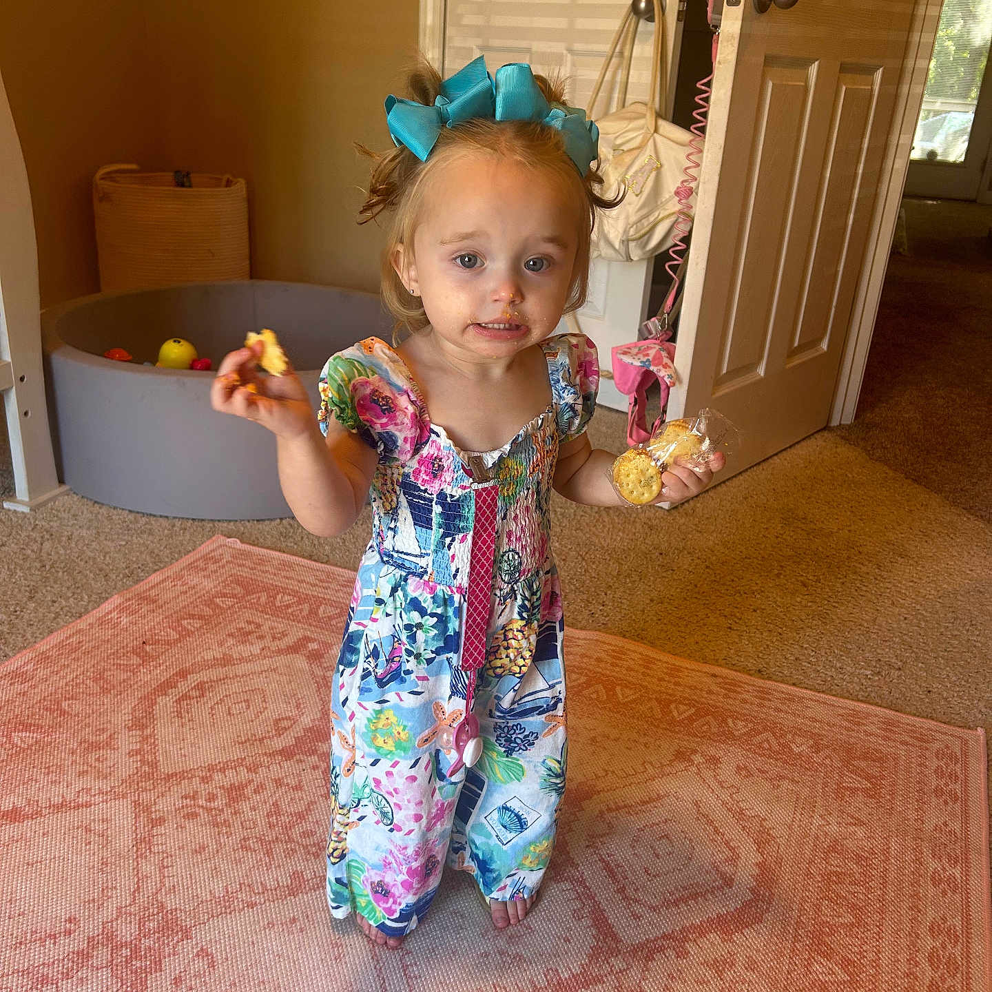 Finlee Blayke is registered to the contest to win money with this photo: blue_bow, carpet, carpeted_floor, child, colorful_clothing, crackers, door, expression, girl, home, indoor, living_room, messy_face, natural_light, play_area, rug, snack, standing, toy_basket, young_child