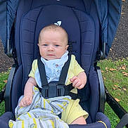 Reggie is registered to the contest to win money with this photo: baby, stroller, outdoor, grass, blanket, yellow_clothing, bib, face, person, infant, baby_boy, blue_stroller, hand, seat_belt, nature, daylight, expression, child, curious, sitting