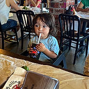 Mateo is registered to the contest to win money with this photo: toddler, child, restaurant, drink, straw, table, napkin_holder, lime, brick_wall, chair, people, casual_clothing, indoor, food, dining, hairstyle, cup, window, background, young_child