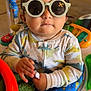 Mateo is registered to the contest to win money with this photo: activity_center, baby, child, clothing, colorful, cute, dinosaur_pajamas, face, hands, head, indoor, infant, person, play, round_glasses, seated, serious_expression, sunglasses, toy, young_child