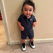 Mateo is registered to the contest to win money with this photo: toddler, child, curly_hair, romper, sneakers, carpet, wall, standing, indoor, looking_up, cute, smiling, young_child, small_hands, shadow, footwear, person, portrait, casual_clothing, curious