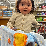 Mateo is registered to the contest to win money with this photo: toddler, child, shopping_cart, blanket, store, shelves, card, clothing, indoor, portrait, curly_hair, face, eyes, expression, retail, aisle, fluorescent_lighting, casual_wear, cute, baby
