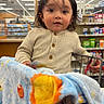 toddler, child, shopping_cart, blanket, store, shelves, card, clothing, indoor, portrait, curly_hair, face, eyes, expression, retail, aisle, fluorescent_lighting, casual_wear, cute, baby