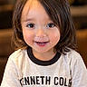 child, toddler, portrait, smiling, happy, face, eyes, teeth, long_hair, brown_hair, t_shirt, clothing, graphic_text, indoor, warm_lighting, close_up, sitting, person, photography, adorable