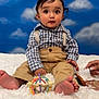 baby, background, barefoot, blue_sky, checkered_shirt, child, clouds, colorful_toy, curly_hair, cute, expression, fluffy_rug, hands, indoor, playful, portrait, seated, suspenders, tan_pants, toy