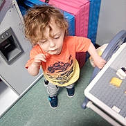 Dominic is registered to the contest to win money with this photo: child, toddler, curly_hair, orange_shirt, indoor, play_area, floor, toy_kitchen, colorful_mats, shoes, motion, young_child, person, play, room, furniture, plastic_toys, casual_clothing, standing, activity