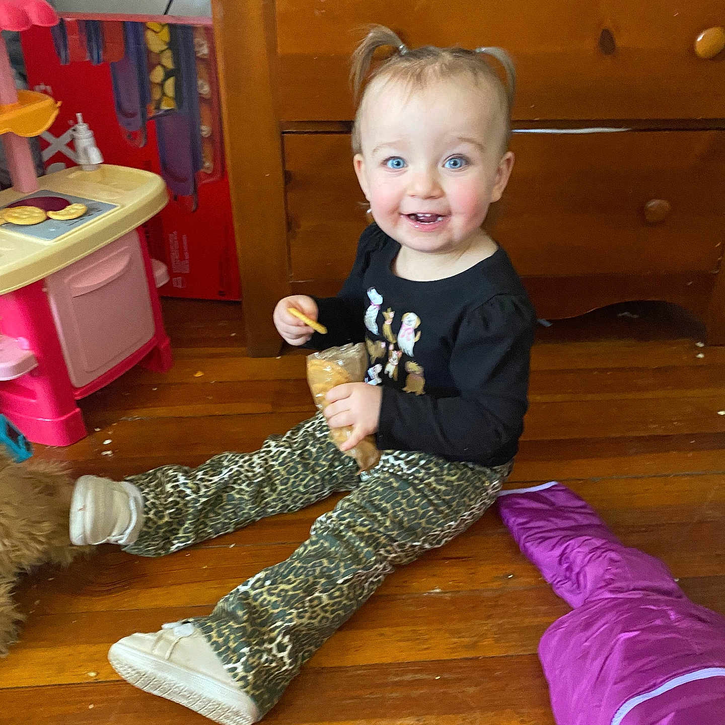 Gianna is registered to the contest to win money with this photo: toddler, child, smiling, indoor, wooden_floor, leopard_print, pants, black_top, pigtails, snack, play_kitchen, purple_sleeping_bag, toy, happy, cute, blue_eyes, girl, casual_clothing, home, floor