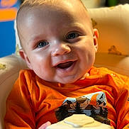 Xander is registered to the contest to win money with this photo: baby, background_blur, blue_eyes, child, close_up, cozy, cup, cute, face, happy, indoor, infant, orange_shirt, person, portrait, sitting, smiling, toy, warm_lighting, young_child