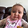 baby, child, pink_bib, elephant_embroidery, food, spoon, leather_couch, face, expression, feeding, clothing, sitting, indoor, cute, chubby_cheeks, person, portrait, young_child, messy, feeding_time