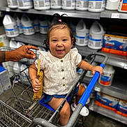 Alani joined the competition — help win amazing prizes! baby, child, shopping_cart, smile, person, store, aisle, bottles, cleaning_supplies, hand, white_clothing, blue_shorts, footwear, woven_bag, hair_tie, happy, indoor, retail, metal_cart, curly_hair